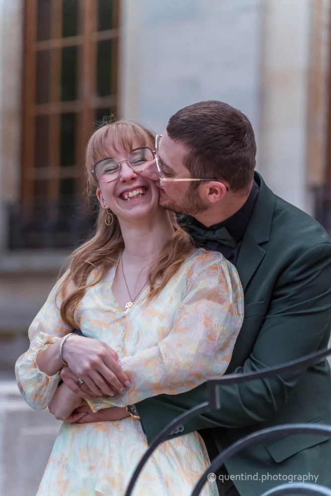 Couple souriant lors de leur seance engagement, femme à lunettes en robe fleurie, homme en costume vert