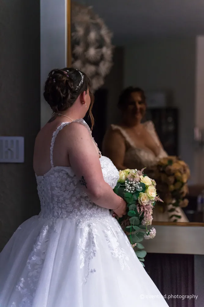 Mariée en préparatifs devant un miroir, tenant son bouquet, robe de dentelle blanche
