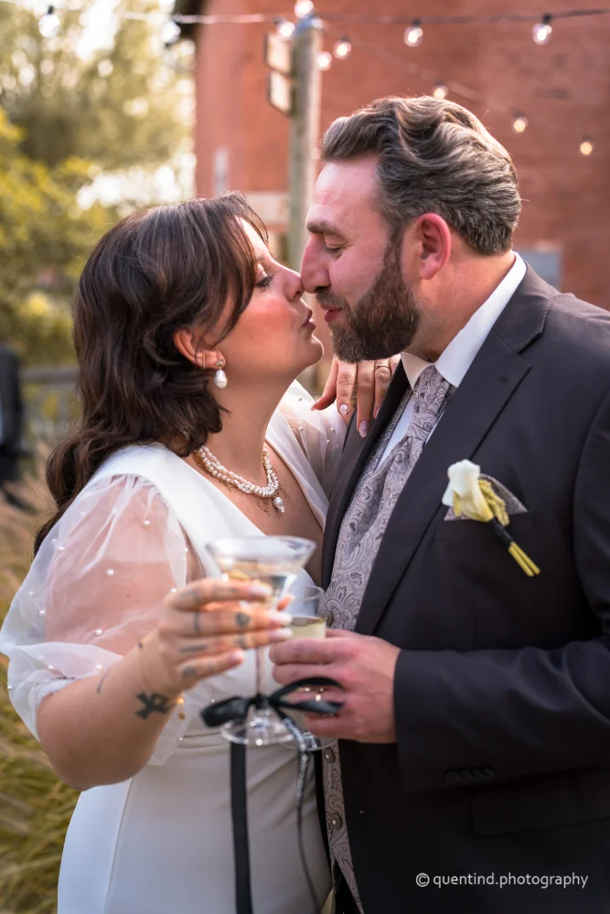 Couple de mariés s'embrassant lors de leur mariage, avec un verre de cocktail à la main et une ambiance romantique sous des guirlandes lumineuses.